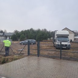Krzysztof Staroń - Pracownik w jaskrawej kurtce montuje siatkę na metalowych słupkach ogrodzenia panelowego, na tle samochód dostawczy i dom jednorodzinny w pochmurny dzień.