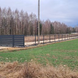 Krzysztof Staroń - Nowoczesne, antracytowe ogrodzenie panelowe z poziomymi prętami, połączone z siatką leśną, otaczające teren z młodym zbożem, widok w kierunku budynku gospodarczego w oddali.