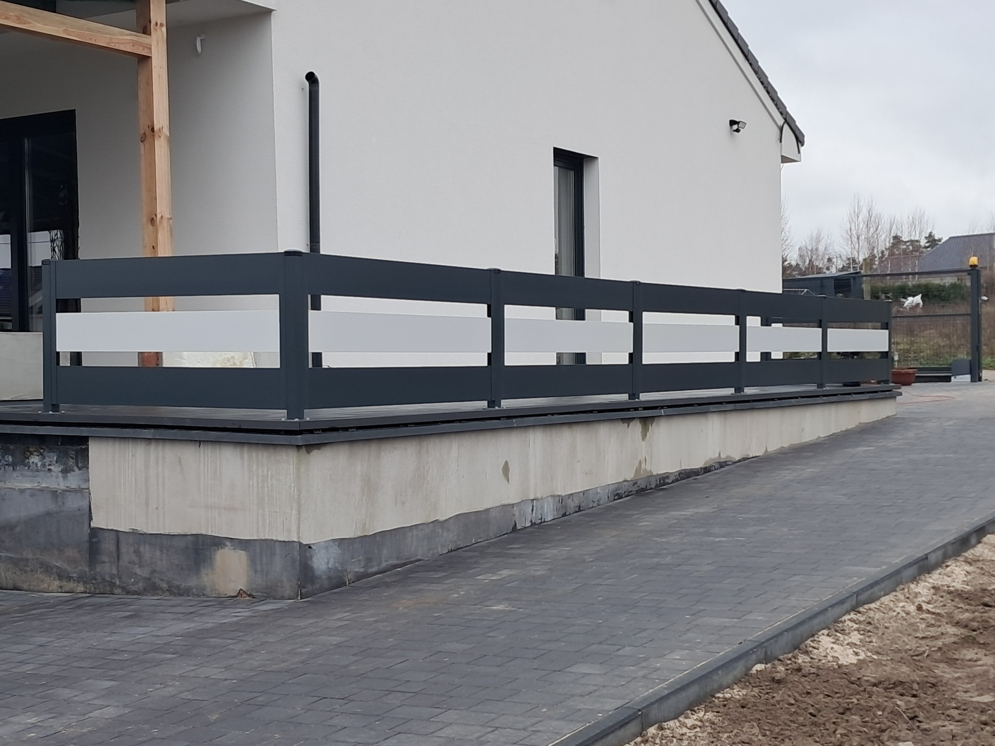 Nowoczesne ogrodzenie panelowe z szarymi ramami i białymi wypełnieniami, zamontowane na podmurówce przy nowym domu z szarą kostką brukową na podjeździe, widok z boku.