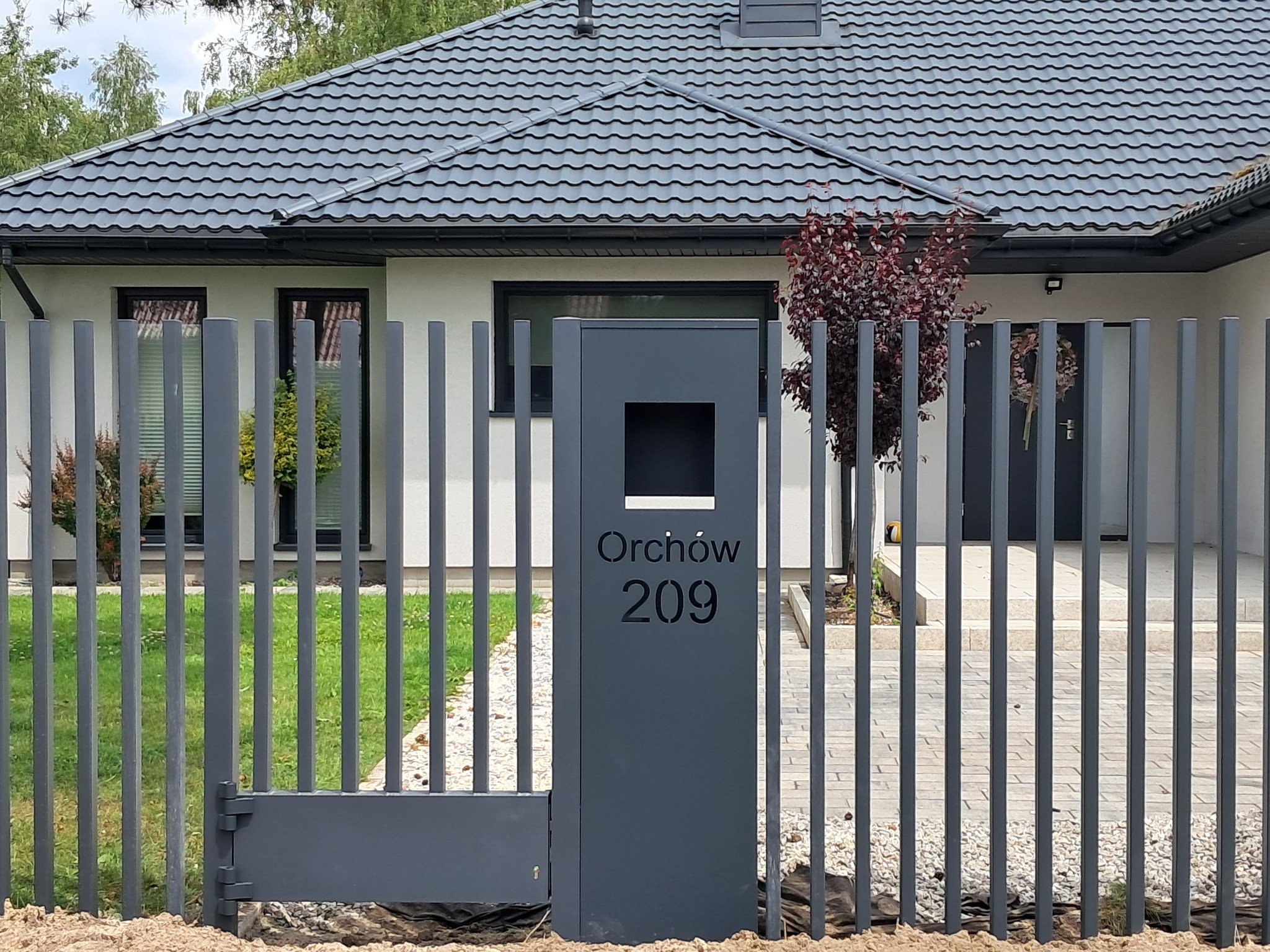 Nowoczesne, grafitowe ogrodzenie z pionowych prętów z bramką i słupkiem z numerem domu 'Orchów 209' na tle jasnego domu z ciemnoszarą dachówką.