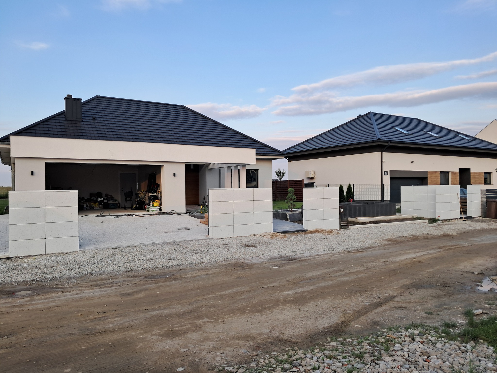 Wznoszenie ogrodzenia z betonowych bloczków modułowych przed nowoczesnymi domami jednorodzinnymi, widoczny plac budowy i materiały.