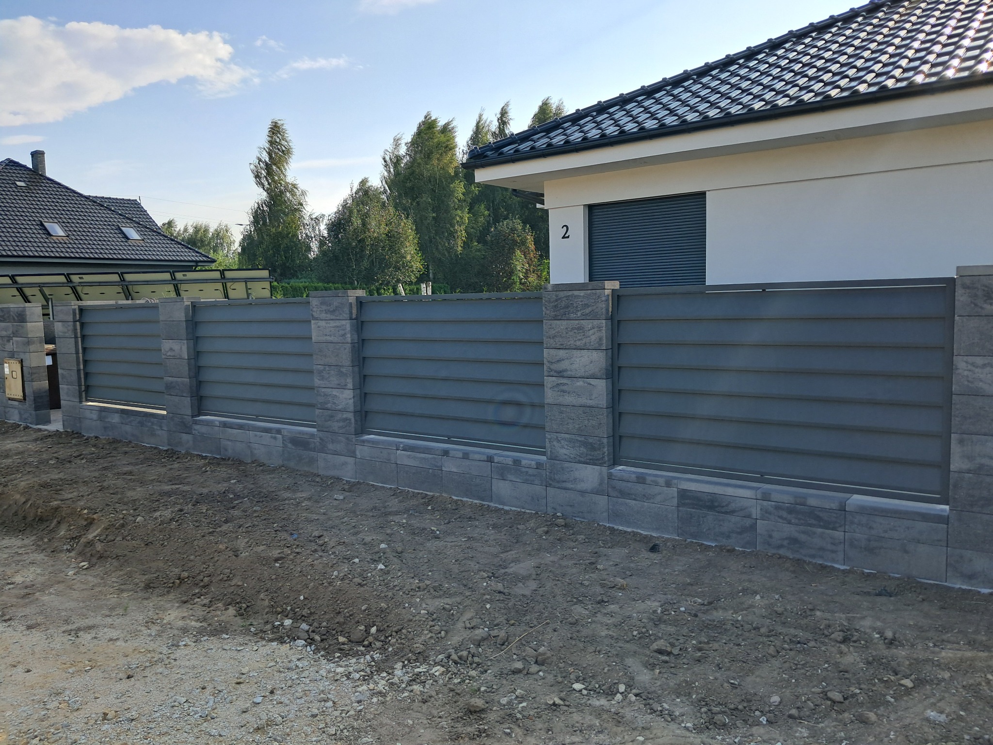 Nowoczesne ogrodzenie z szarych paneli horyzontalnych i kamiennych słupków na tle domu z ciemną dachówką i zielenią.