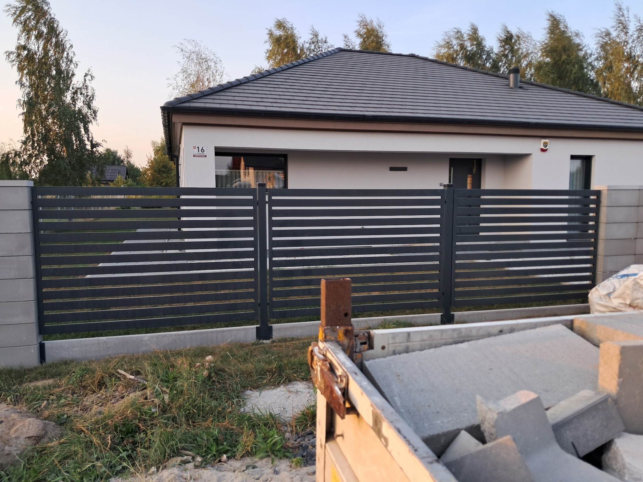 Nowoczesne, poziome przęsła ogrodzeniowe w kolorze grafitowym, zamontowane na betonowych słupkach, z widocznym fragmentem domu z szarą dachówką i elementami transportowymi w tle.