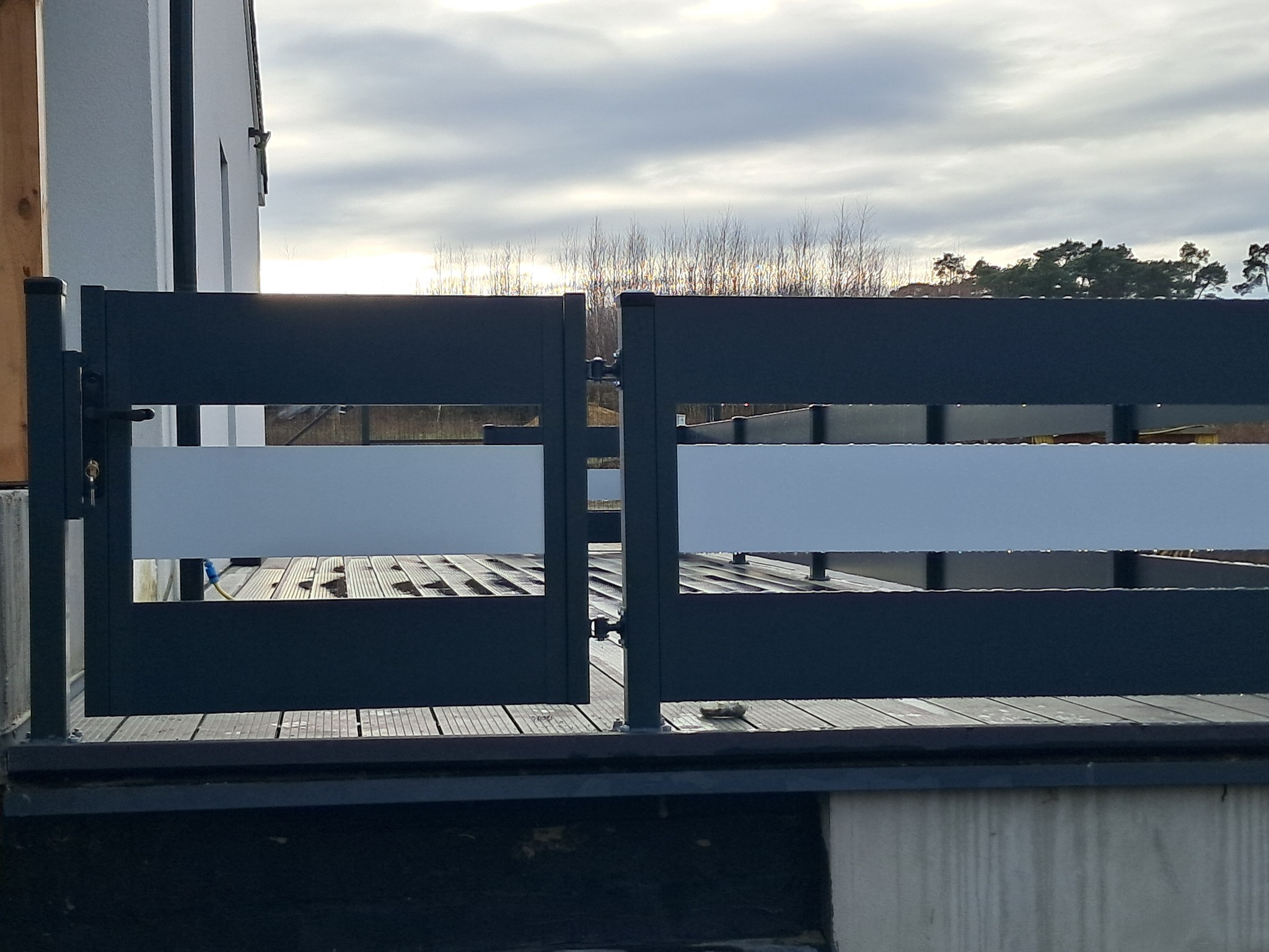 Nowoczesna, otwarta brama z poziomymi panelami w kolorze grafitowym, z widocznym drewnianym podestem i zabudową domu w tle, oświetlona zachodzącym słońcem.