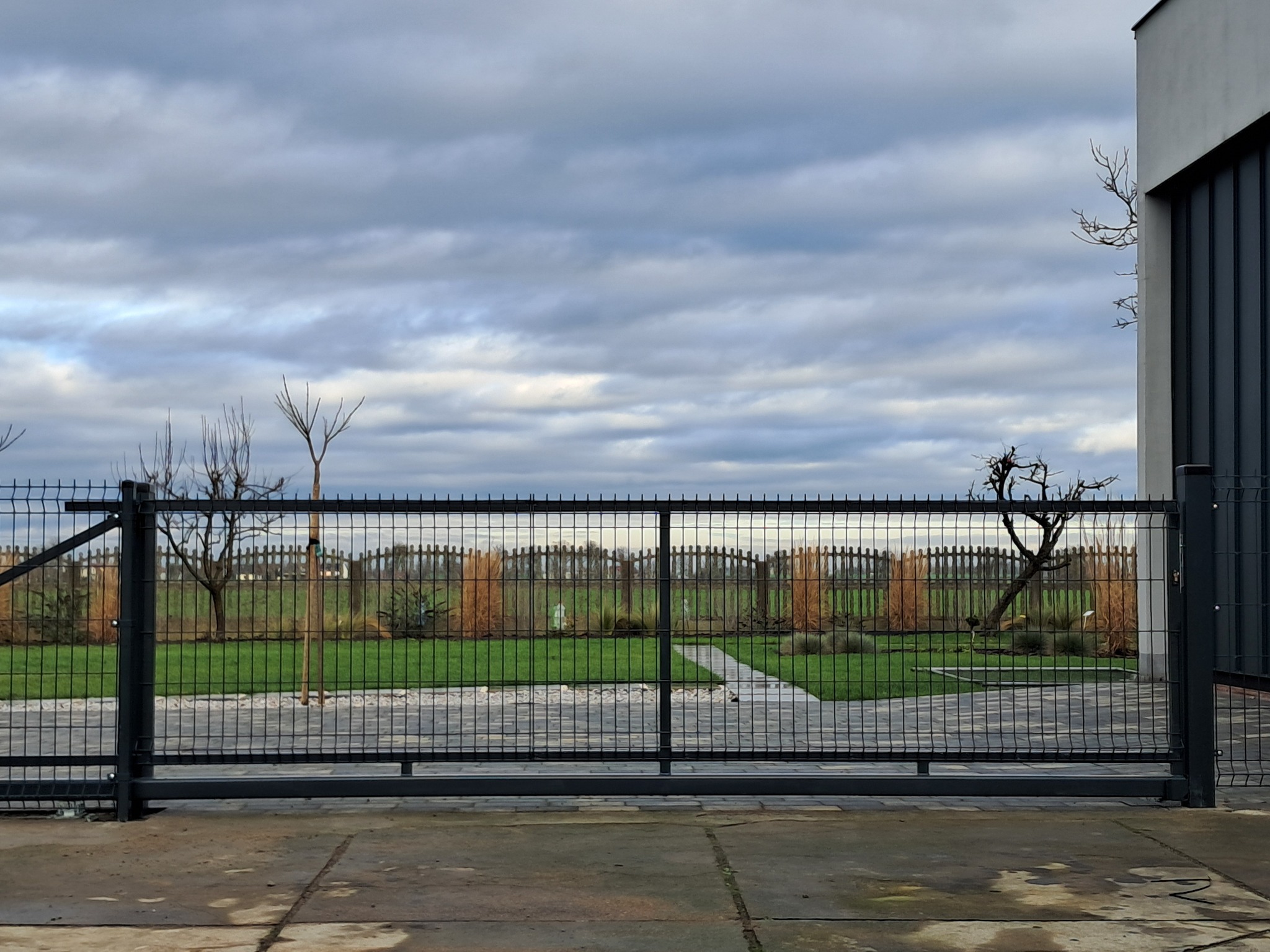 Przesuwna brama z czarnej siatki na tle zadbanego ogrodu z widocznym fragmentem nowoczesnego domu i pochmurnym niebem.