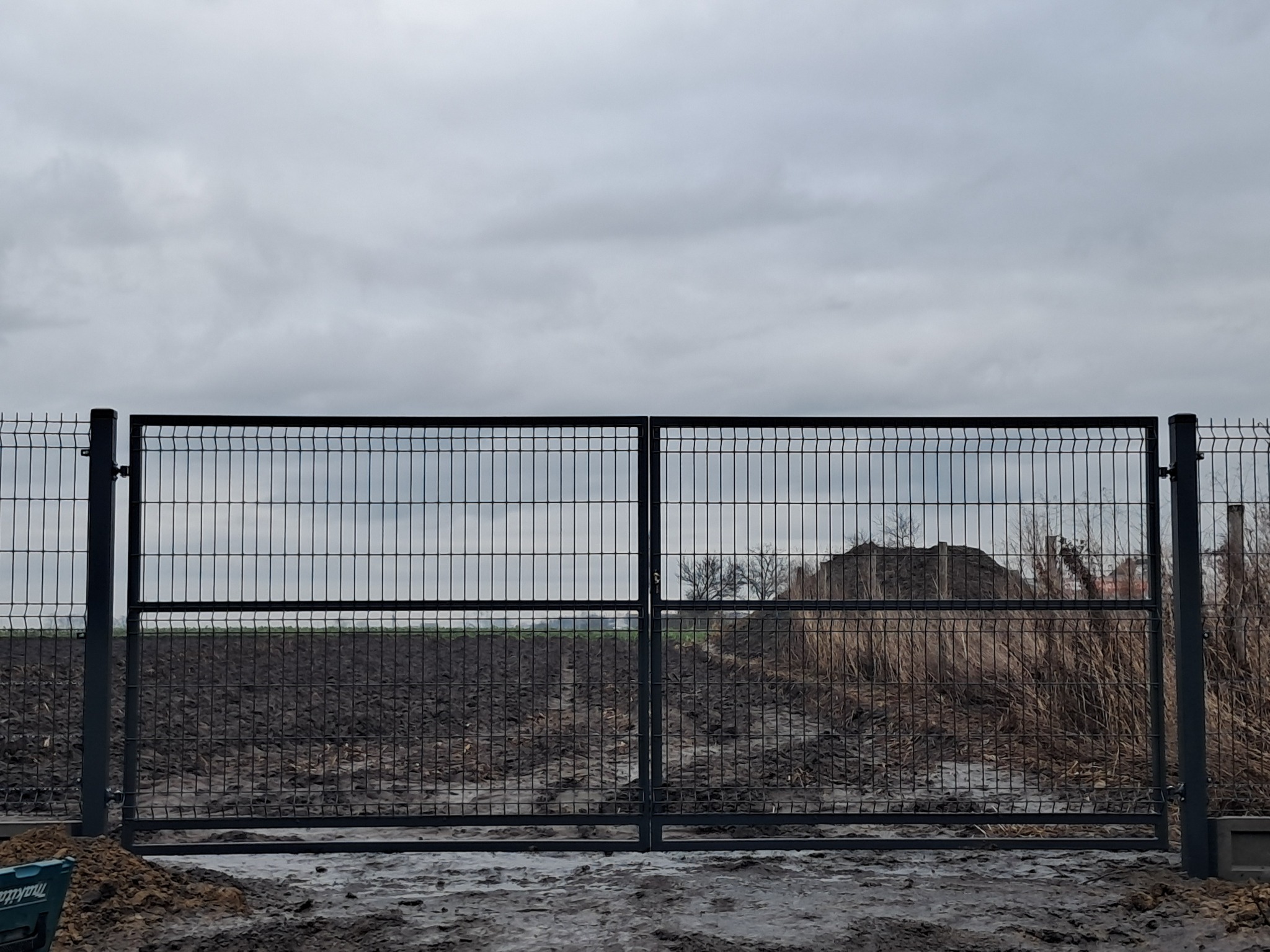 Zamknięta, metalowa brama dwuskrzydłowa z siatki zgrzewanej, widoczna na tle zaoranego pola i pochmurnego nieba. W lewym dolnym rogu widoczny fragment niebieskiej skrzynki narzędziowej.