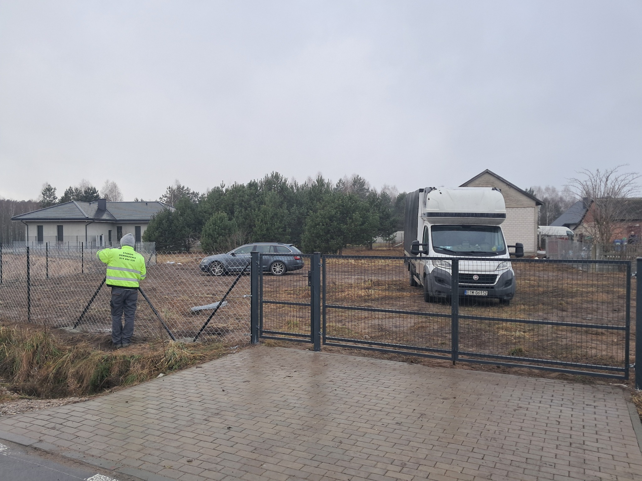 Pracownik w jaskrawej kurtce montuje siatkę na metalowych słupkach ogrodzenia panelowego, na tle samochód dostawczy i dom jednorodzinny w pochmurny dzień.