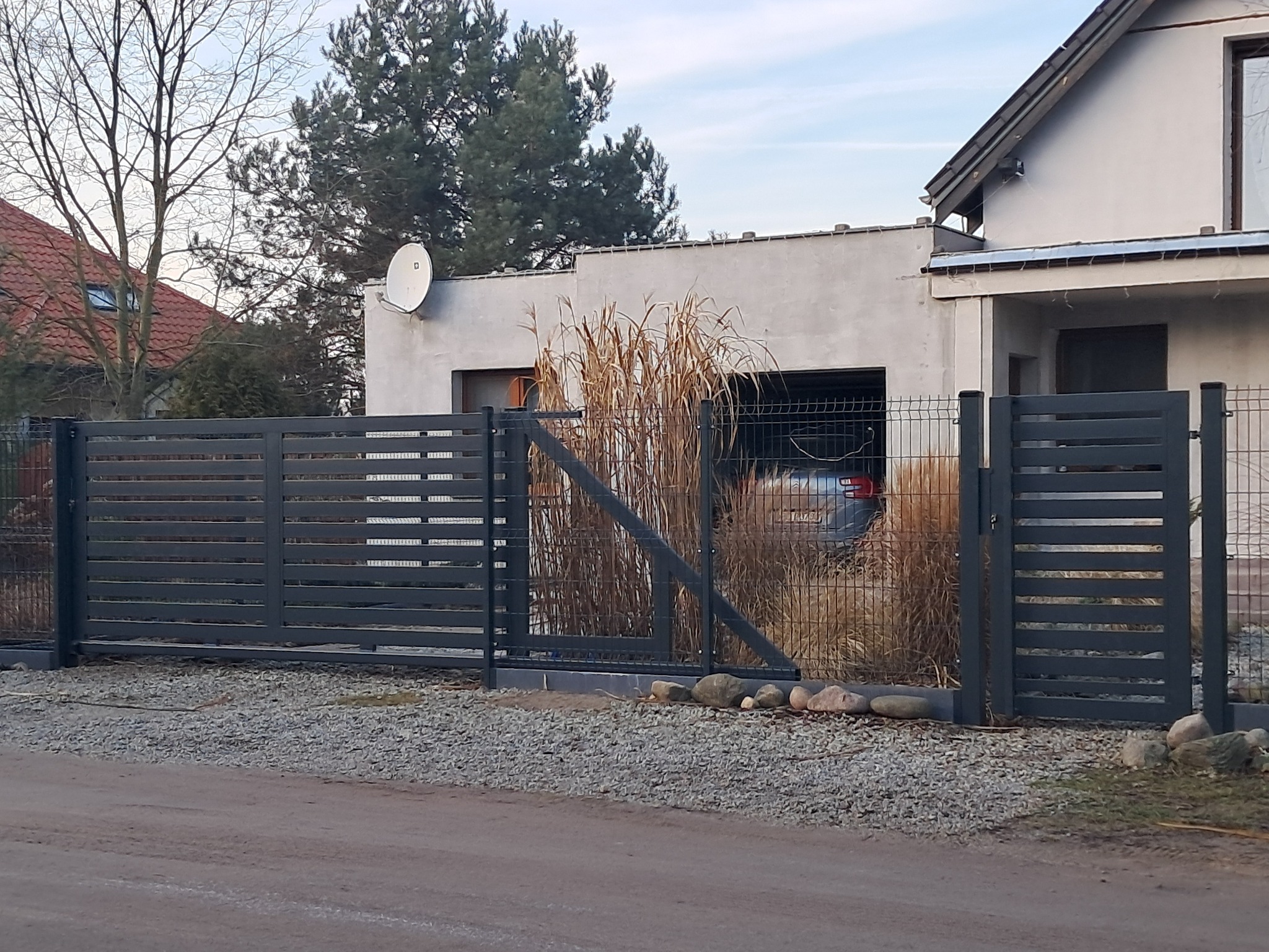 Nowoczesna, pozioma brama wjazdowa i furtka w kolorze grafitowym, z widoczną siatką ogrodzeniową w tle i samochodem w garażu, otoczone ozdobnymi trawami i kamieniami.