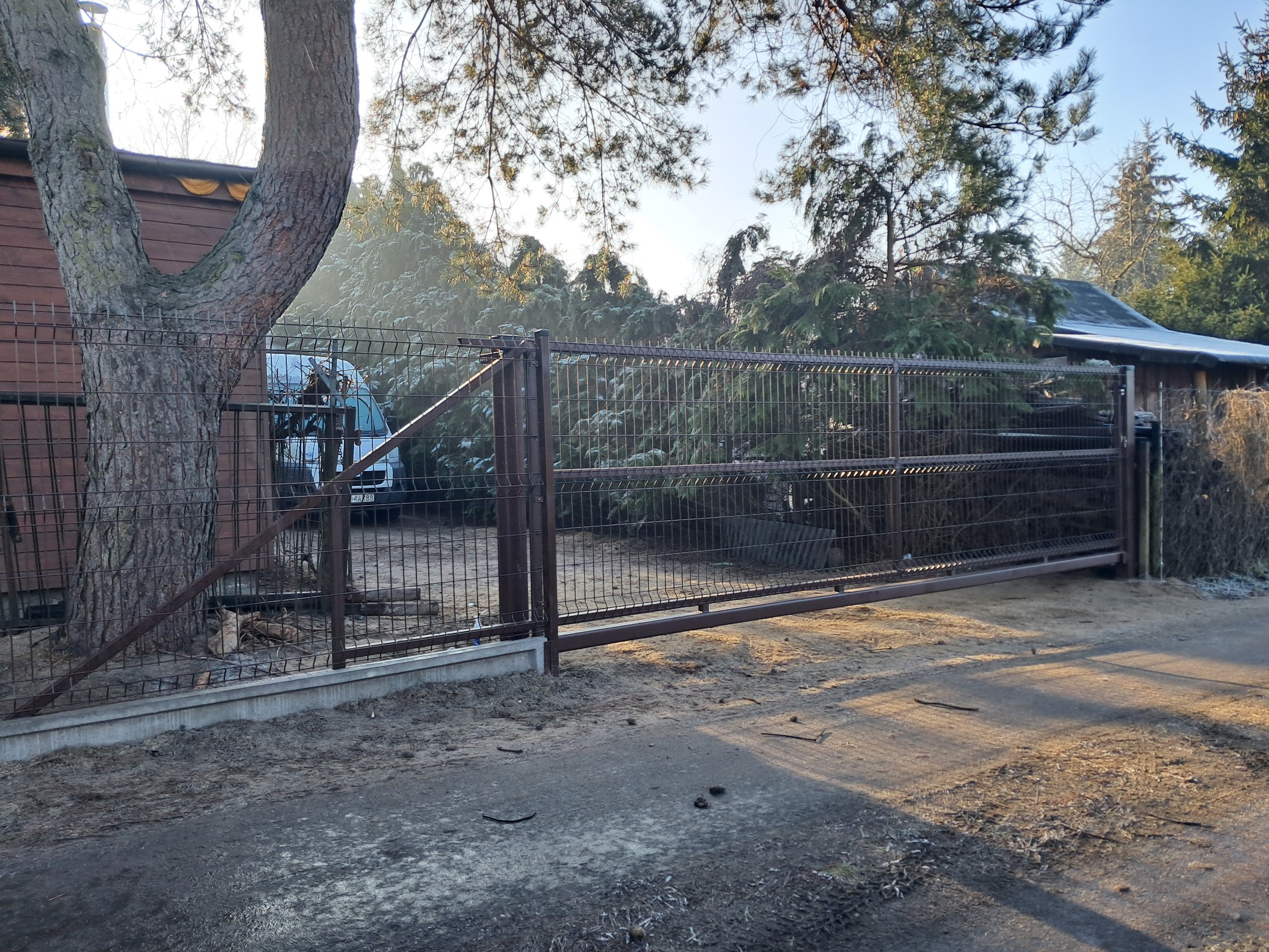 Zamykana brama w metalowym ogrodzeniu panelowym z betonową podmurówką, widok z zewnątrz posesji w słoneczny dzień, w tle zabudowania i zieleń.