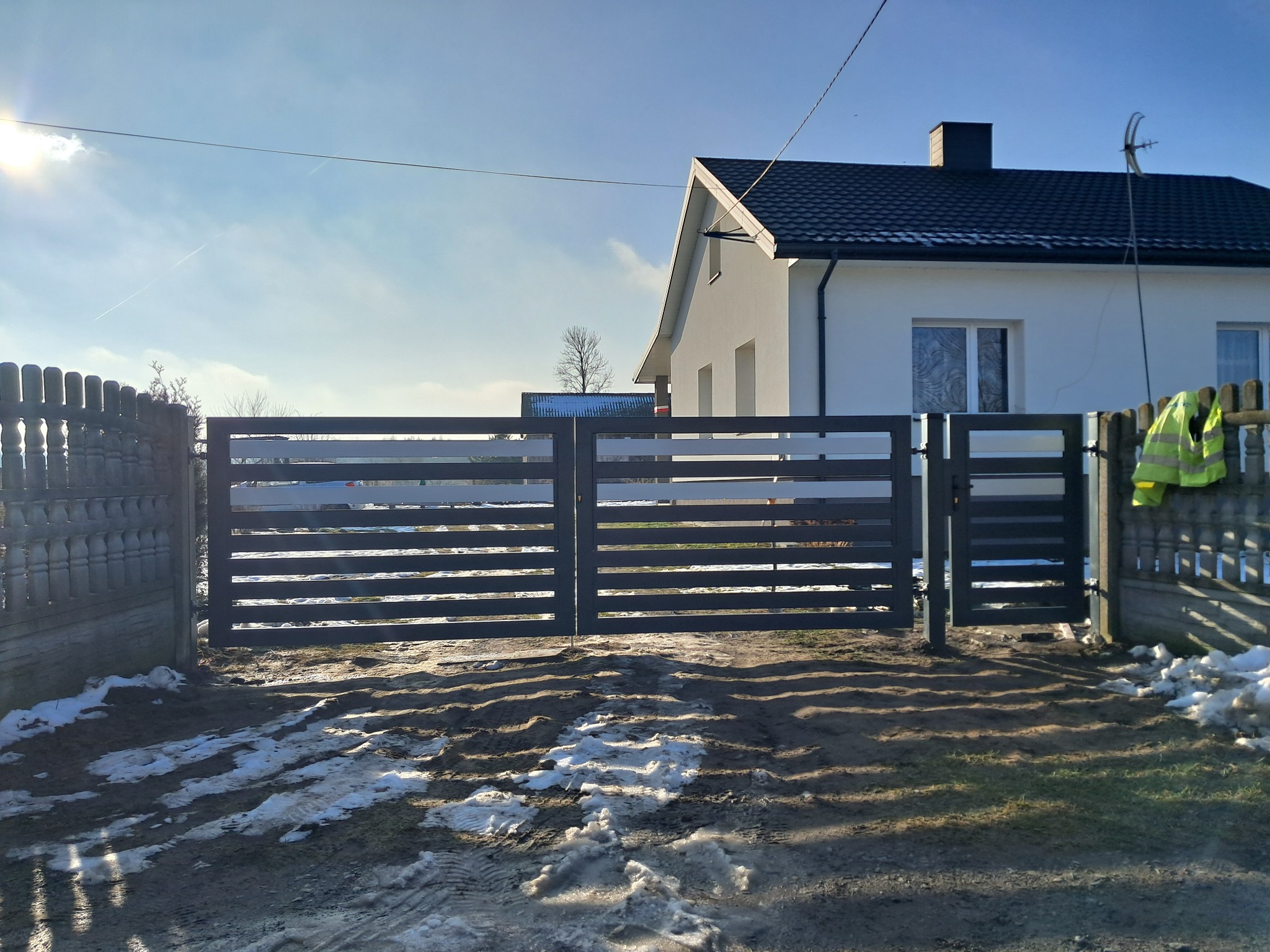 Nowoczesna, pozioma brama wjazdowa w kolorze antracytowym, z widocznym domem w tle i resztkami śniegu na ziemi, oświetlona jasnym słońcem.