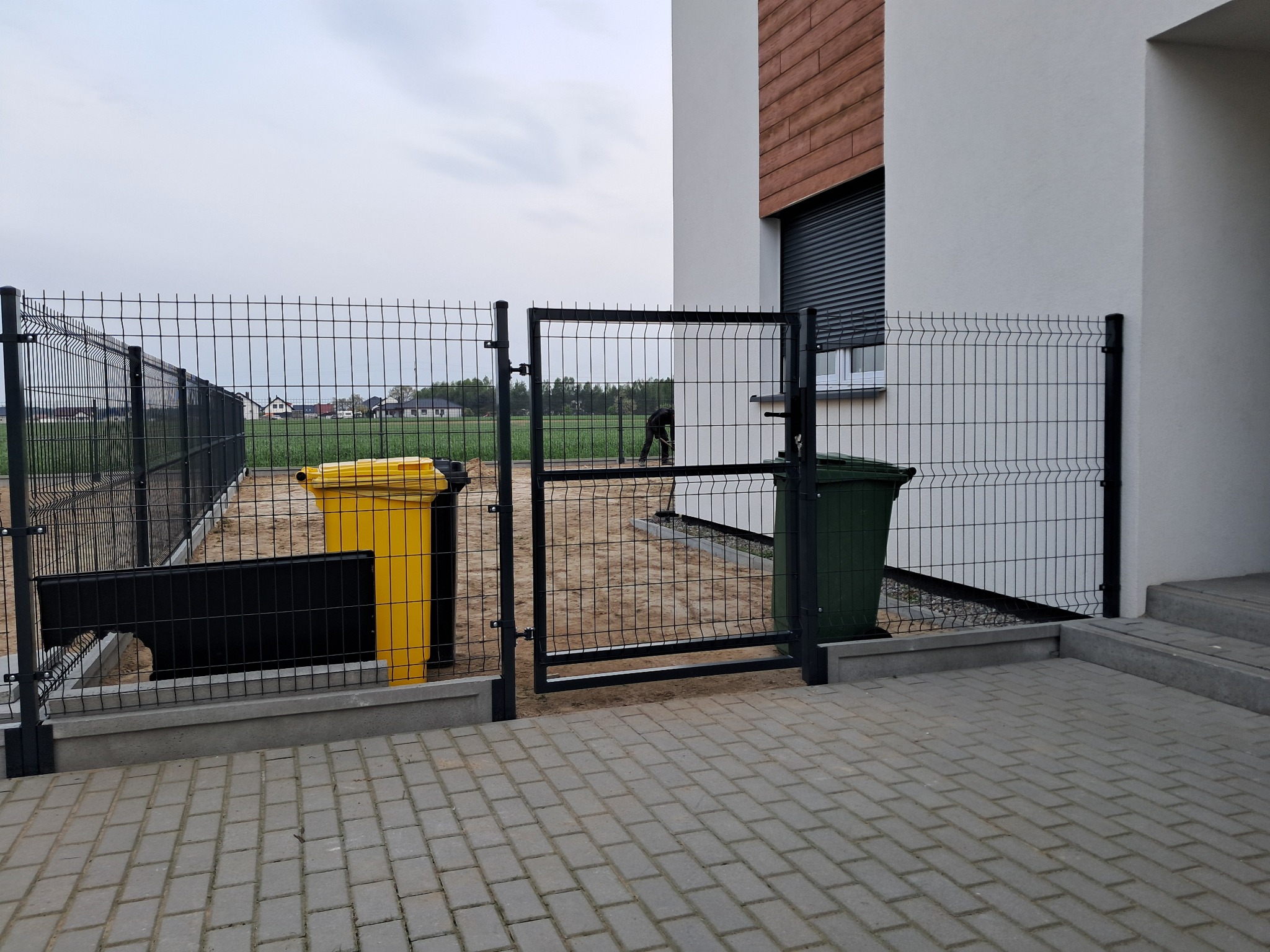 Czarna brama i ogrodzenie panelowe z widocznymi żółtym i zielonym pojemnikiem na śmieci, na tle częściowo wykończonego domu z drewnianą okładziną.