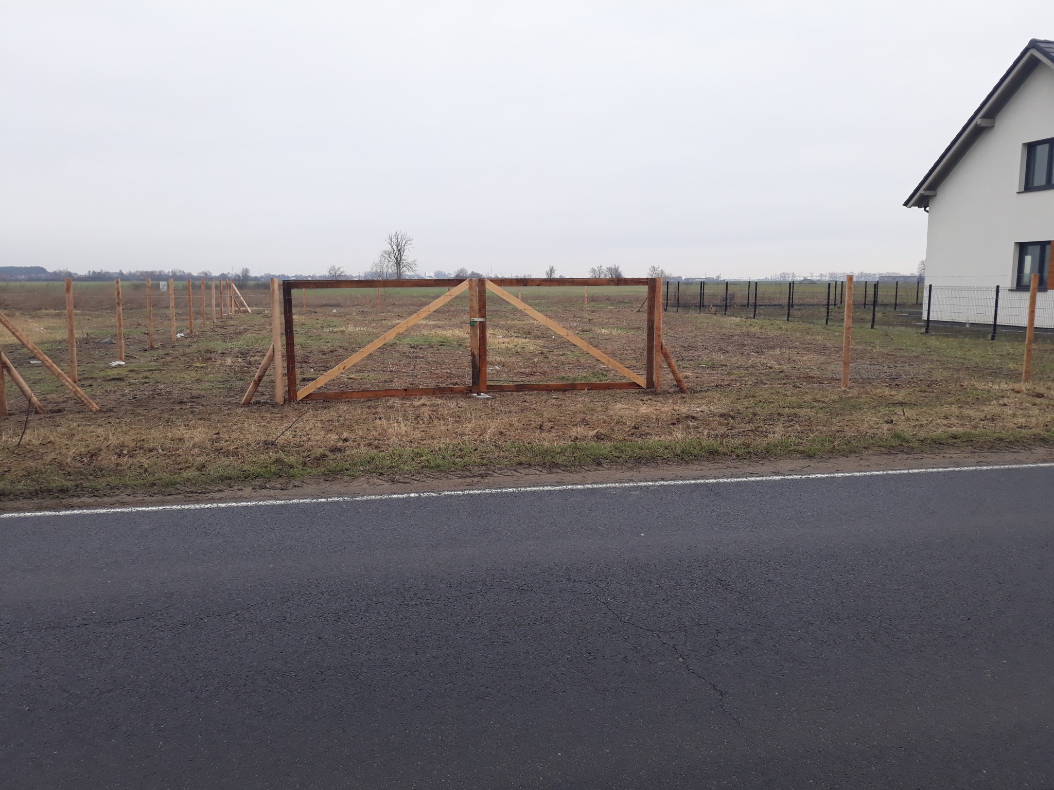 Wstępny etap budowy ogrodzenia z drewnianymi słupkami i ramą bramy na tle domu i pola w pochmurny dzień.