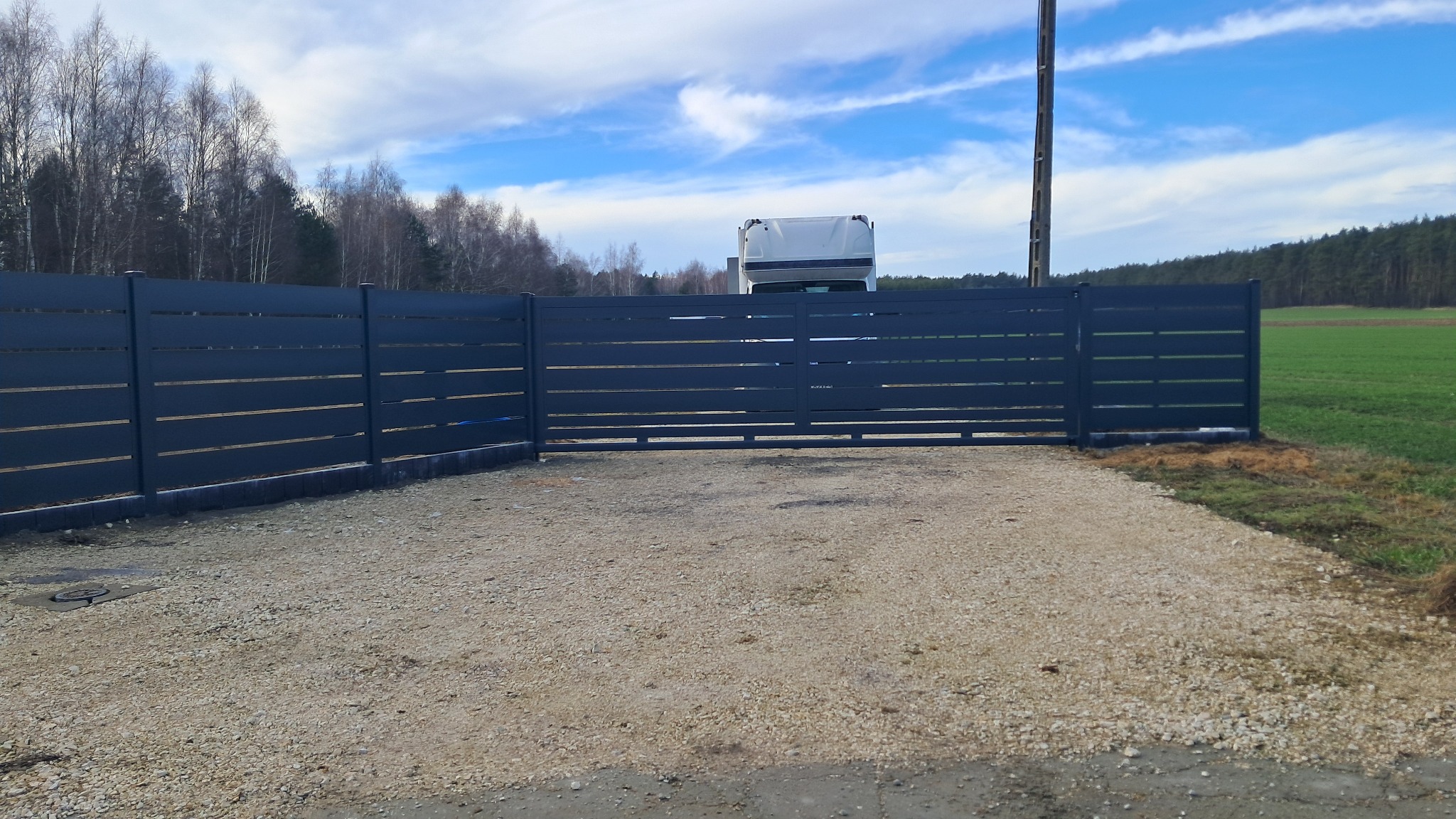 Nowoczesne, poziome, szare ogrodzenie z metalowymi przęsłami i bramą wjazdową na żwirowym podłożu, z widocznym fragmentem białej ciężarówki w tle.