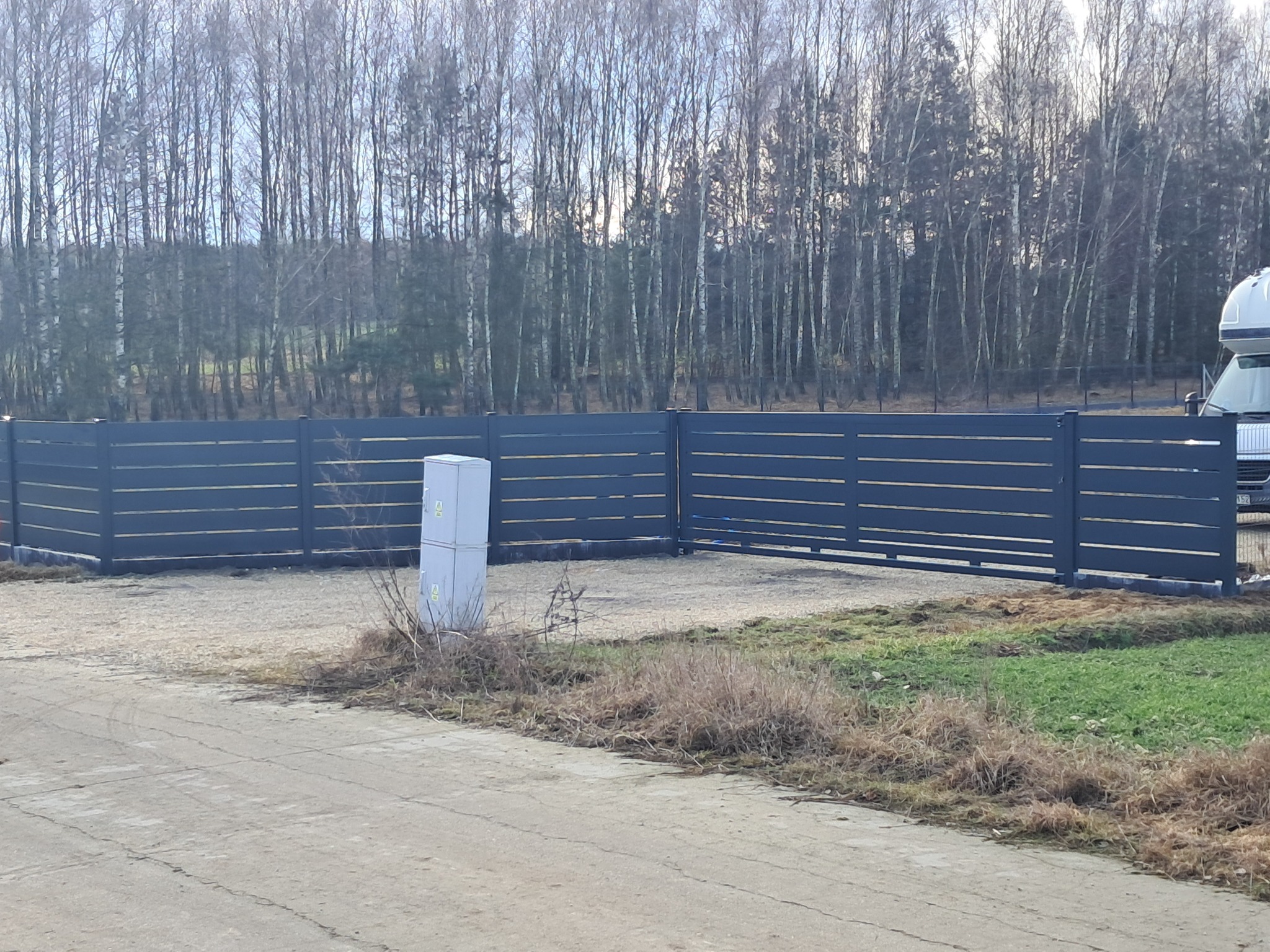 Nowoczesne ogrodzenie metalowe z poziomych paneli w kolorze grafitowym, z bramą wjazdową, na tle lasu brzozowego i zaparkowanego kampera.