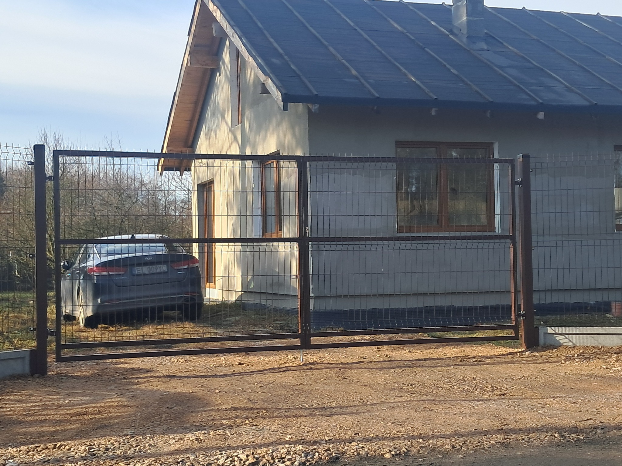 Nowoczesna brama z siatki metalowej w kolorze brązowym, z widocznym samochodem osobowym i fragmentem domu w tle.