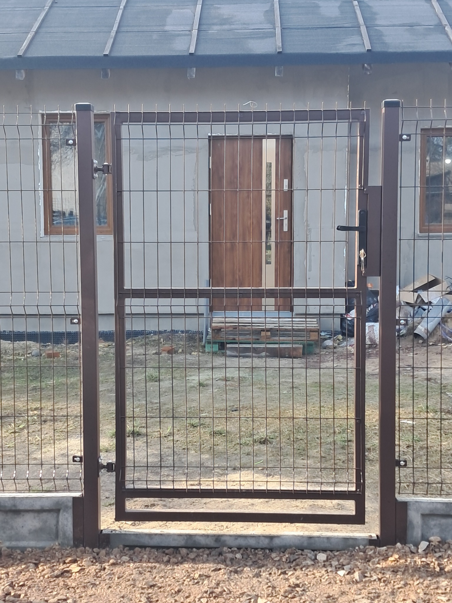 Brązowa furtka z metalowej siatki na betonowej podmurówce, prowadząca do domu w trakcie budowy, widok z zewnątrz.