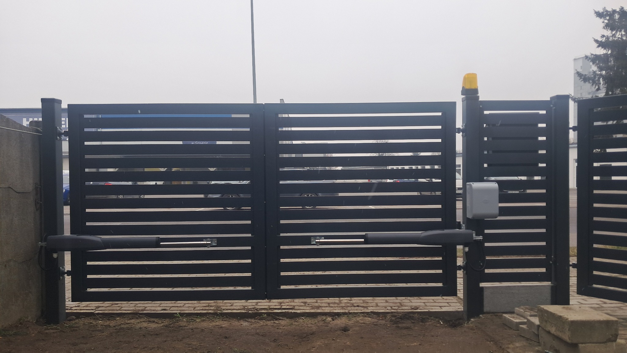 Automatyczna, pozioma brama metalowa z napędem w Jasinie, widok z zewnątrz, z żółtą lampą ostrzegawczą i szarym sterownikiem.