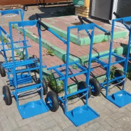 Trzy niebieskie wózki transportowe do butli gazowych, z łańcuchami zabezpieczającymi, stoją na kostce brukowej przed budynkiem z zielonymi akcentami i butlą gazową w tle.