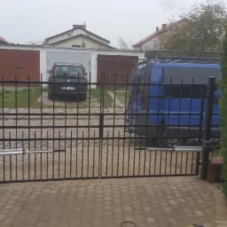 Czarna, metalowa brama wjazdowa z automatyką, widziana z zewnątrz posesji, na tle garaży i zaparkowanych samochodów w pochmurny dzień.