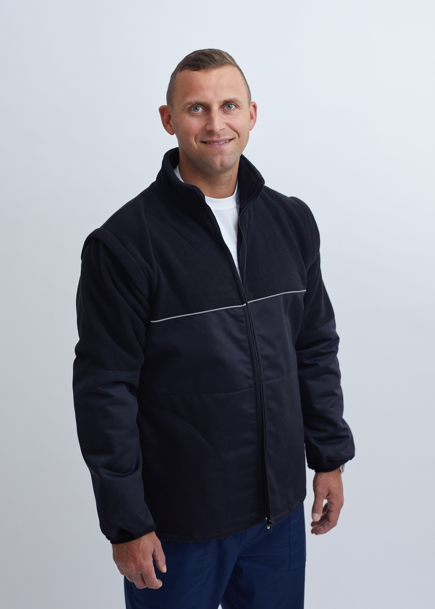 Uśmiechnięty mężczyzna prezentuje ciemną kurtkę roboczą z polarowym kołnierzem i odblaskowym paskiem na piersi, na białym tle.