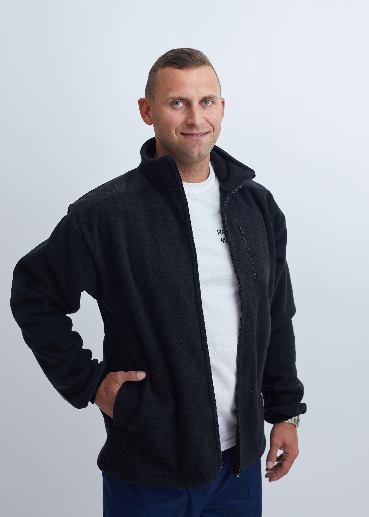 Uśmiechnięty mężczyzna w czarnej polarowej bluzie z suwakiem i kieszenią na piersi, ubrany w białą koszulkę i granatowe spodnie, pozuje na jasnym tle.