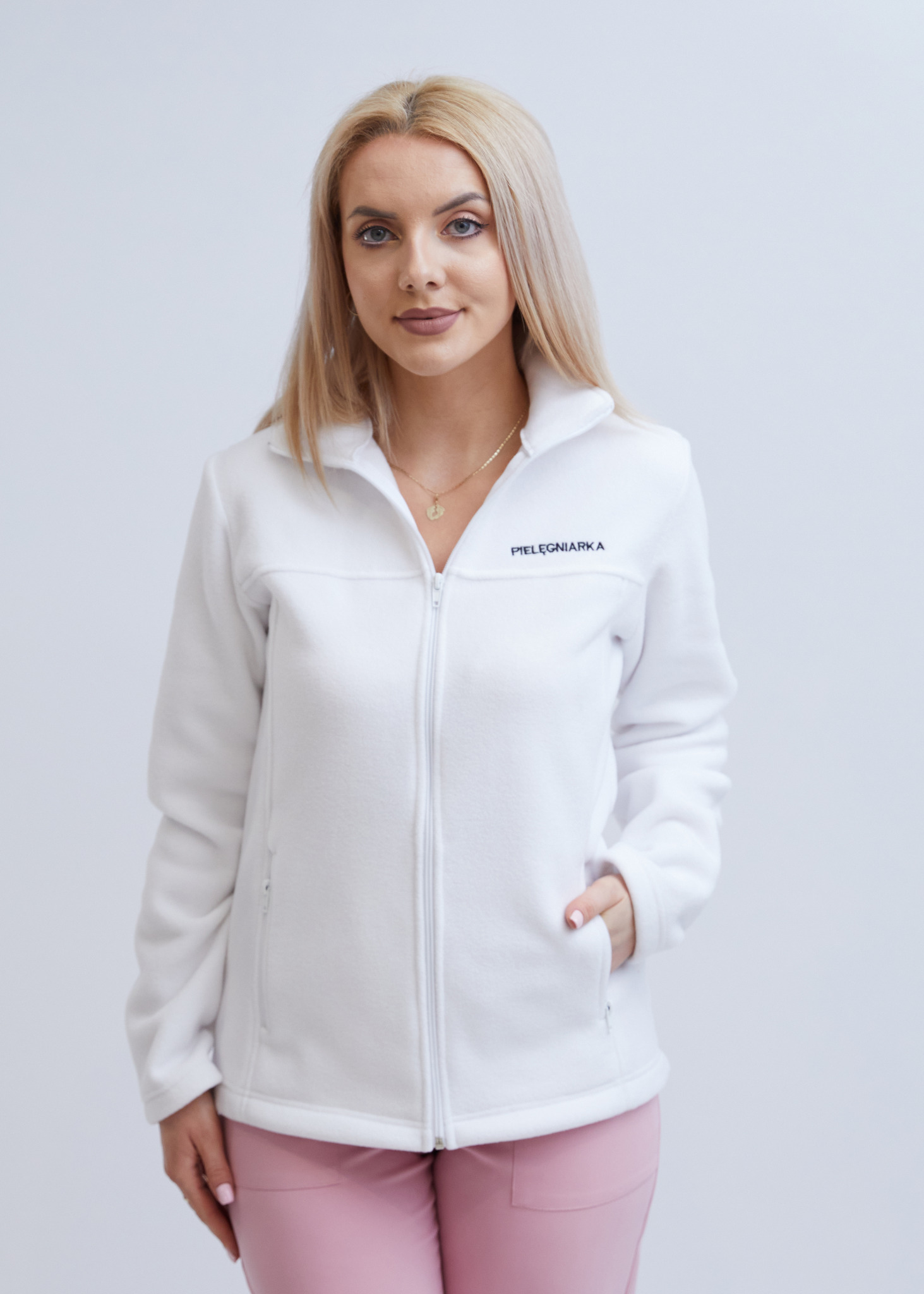 Blondwłosa kobieta w białej polarowej bluzie medycznej z napisem 'PIELĘGNIARKA' i różowych spodniach na jednolitym, jasnym tle.