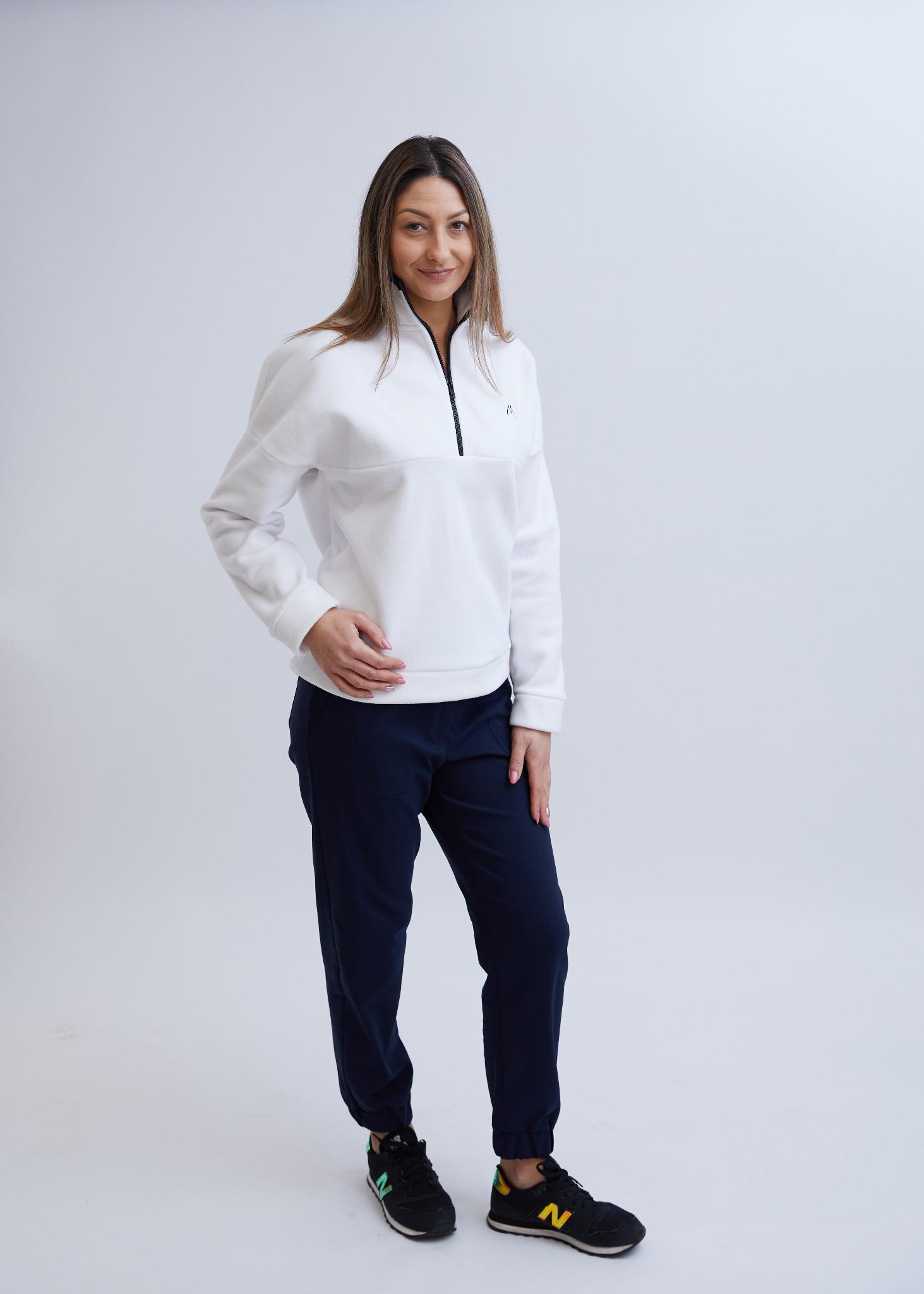 Kobieta w białej bluzie z krótkim zamkiem i granatowych spodniach dresowych, pozująca na jasnym tle.