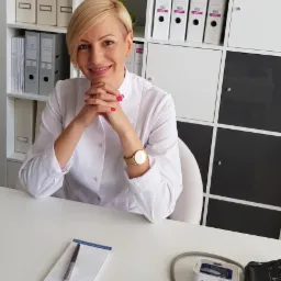 Uśmiechnięta kobieta w białym fartuchu lekarskim siedzi za biurkiem z notatnikiem, długopisem i ciśnieniomierzem, w tle białe regały z segregatorami.