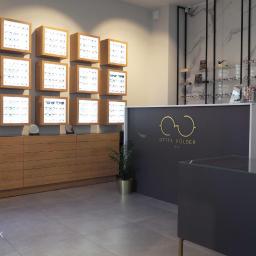 Mazak Studio - Wnętrze salonu optycznego: drewniana komoda z podświetlanymi gablotami na okulary, szary lada recepcyjna z logo 'OPTYK KOLDER 1960', marmurowa ściana z półkami na akcesoria.