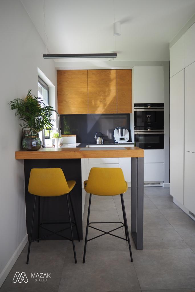 Nowoczesna kuchnia z drewnianymi frontami szafek, blatem z rośliną w szklanym wazonie i dwoma żółtymi krzesłami barowymi na czarnych nogach.