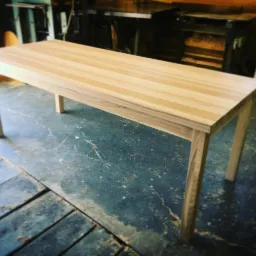 Surowy, prostokątny stół z jasnego drewna w warsztacie, widoczne niedoskonałości podłogi i sprzęt stolarski w tle.