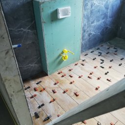 EMIL - Łazienka w trakcie remontu: ułożone płytki podłogowe imitujące drewno, z użyciem klipsów poziomujących, zabudowa stelaża WC z wyprowadzonymi przyłączami, ściany wyłożone szarymi płytkami...