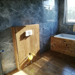 EMIL - Łazienka w trakcie wykańczania: drewnopodobne płytki na podłodze i obudowie wanny, szare płytki imitujące marmur na ścianach, widoczne mocowania do baterii podtynkowych i puszka elektryczna.