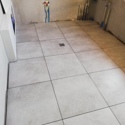 EMIL - Wnętrze łazienki w trakcie remontu, widoczne ułożone jasne płytki podłogowe, instalacja wodna i kanalizacyjna na ścianie, surowy beton. Odpływ liniowy w podłodze.