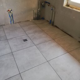 EMIL - Surowa łazienka w trakcie remontu z ułożonymi jasnymi płytkami podłogowymi, widoczne wyprowadzenia instalacji wodno-kanalizacyjnych na ścianach i kratka ściekowa w podłodze.