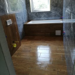 EMIL - Wykończona łazienka z drewnopodobnymi płytkami na podłodze i obudowie wanny, szare marmurowe płytki na ścianach, okno w tle.