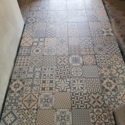 EMIL - Ułożona podłoga z patchworkowych płytek ceramicznych w odcieniach szarości, beżu i brązu, widok z góry na korytarz w trakcie remontu.