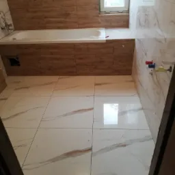 Wykończona łazienka z wanną obudowaną panelami imitującymi drewno, białe marmurowe płytki na ścianach i podłodze, widoczne przyłącza wodne i okno z widokiem na zieleń.