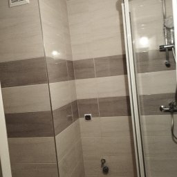 EMIL - Wykończona łazienka z kabiną prysznicową, jasnymi i brązowymi płytkami ułożonymi w poziome pasy, widoczna kratka wentylacyjna i bateria prysznicowa.