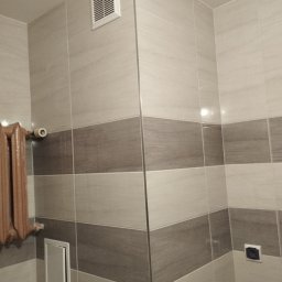 EMIL - Wykończona łazienka z beżowymi i brązowymi płytkami, grzejnikiem żeberkowym, kratką wentylacyjną oraz małą klapką rewizyjną.