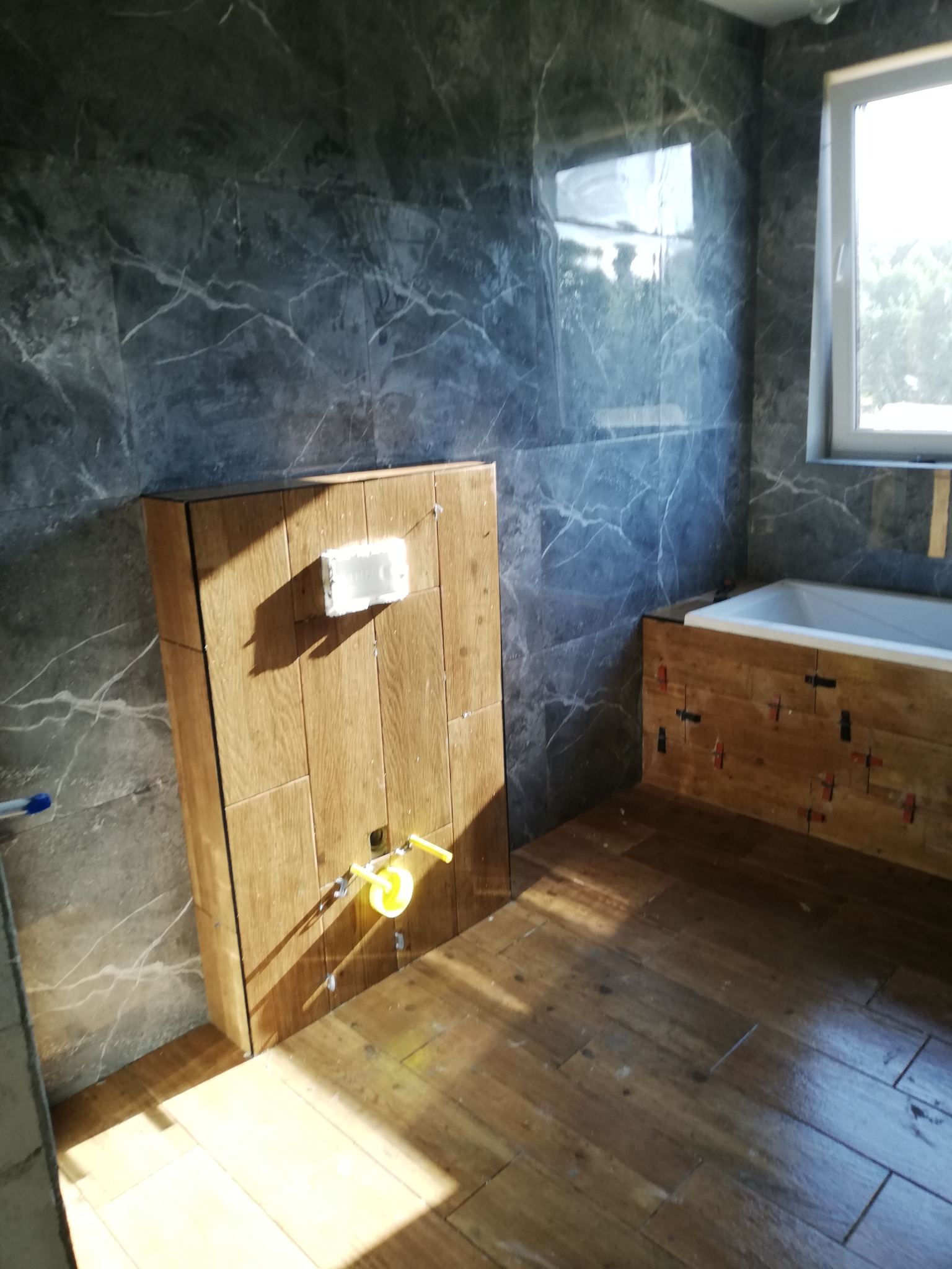 Łazienka w trakcie wykańczania: drewnopodobne płytki na podłodze i obudowie wanny, szare płytki imitujące marmur na ścianach, widoczne mocowania do baterii podtynkowych i puszka elektryczna.