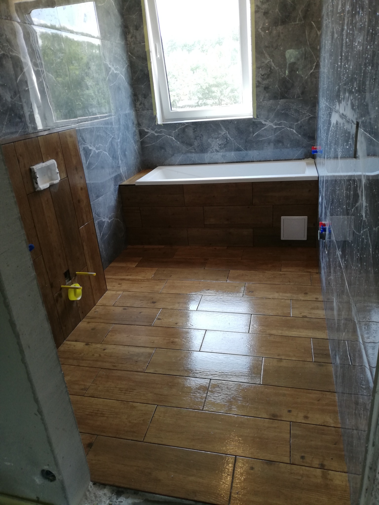 Wykończona łazienka z drewnopodobnymi płytkami na podłodze i obudowie wanny, szare marmurowe płytki na ścianach, okno w tle.