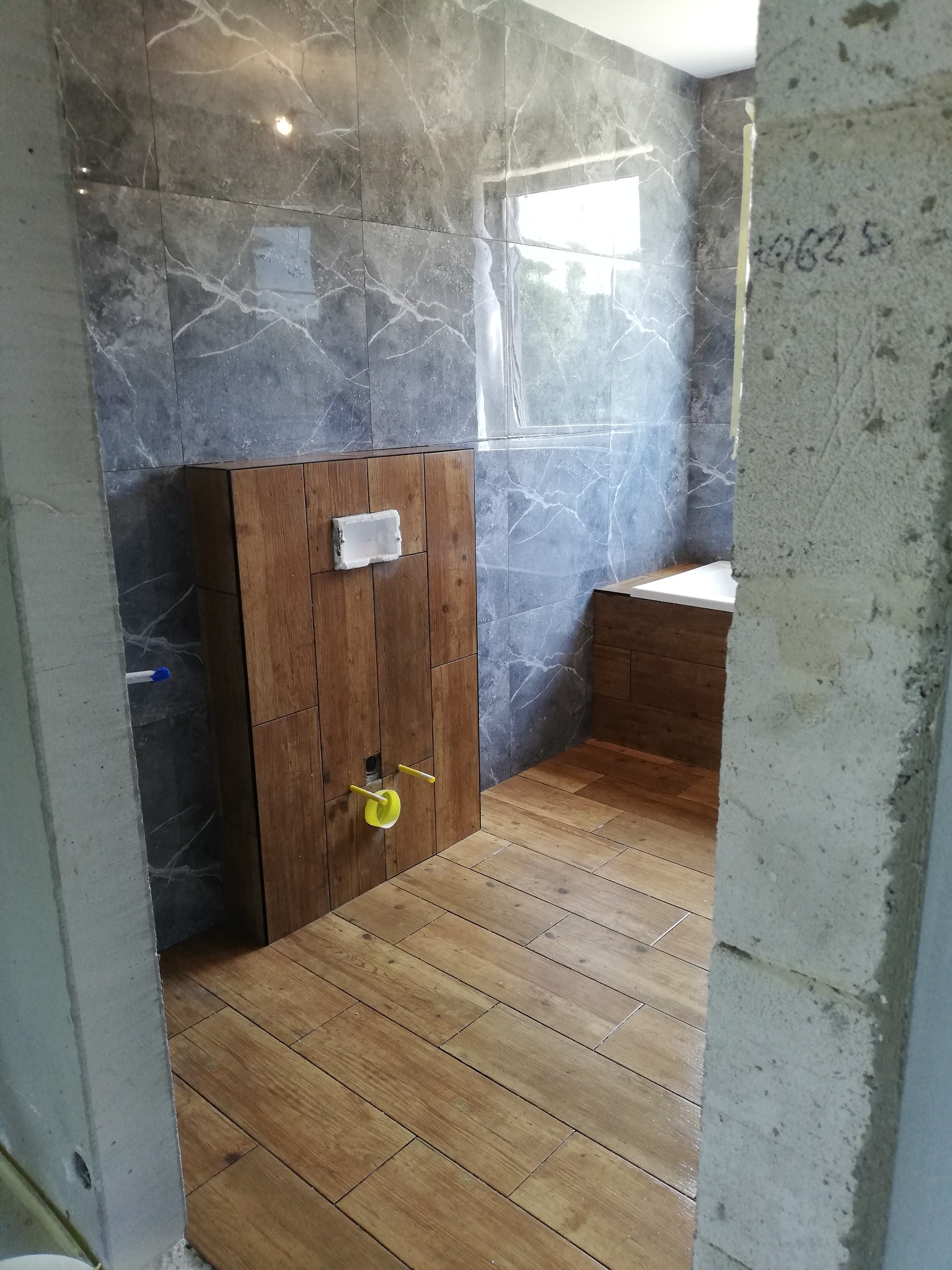 Wnętrze łazienki w trakcie remontu, widoczne szare marmurowe płytki na ścianach, drewnopodobne płytki na podłodze, zabudowa stelaża WC z drewnianymi panelami oraz surowy betonowy filar z napisem