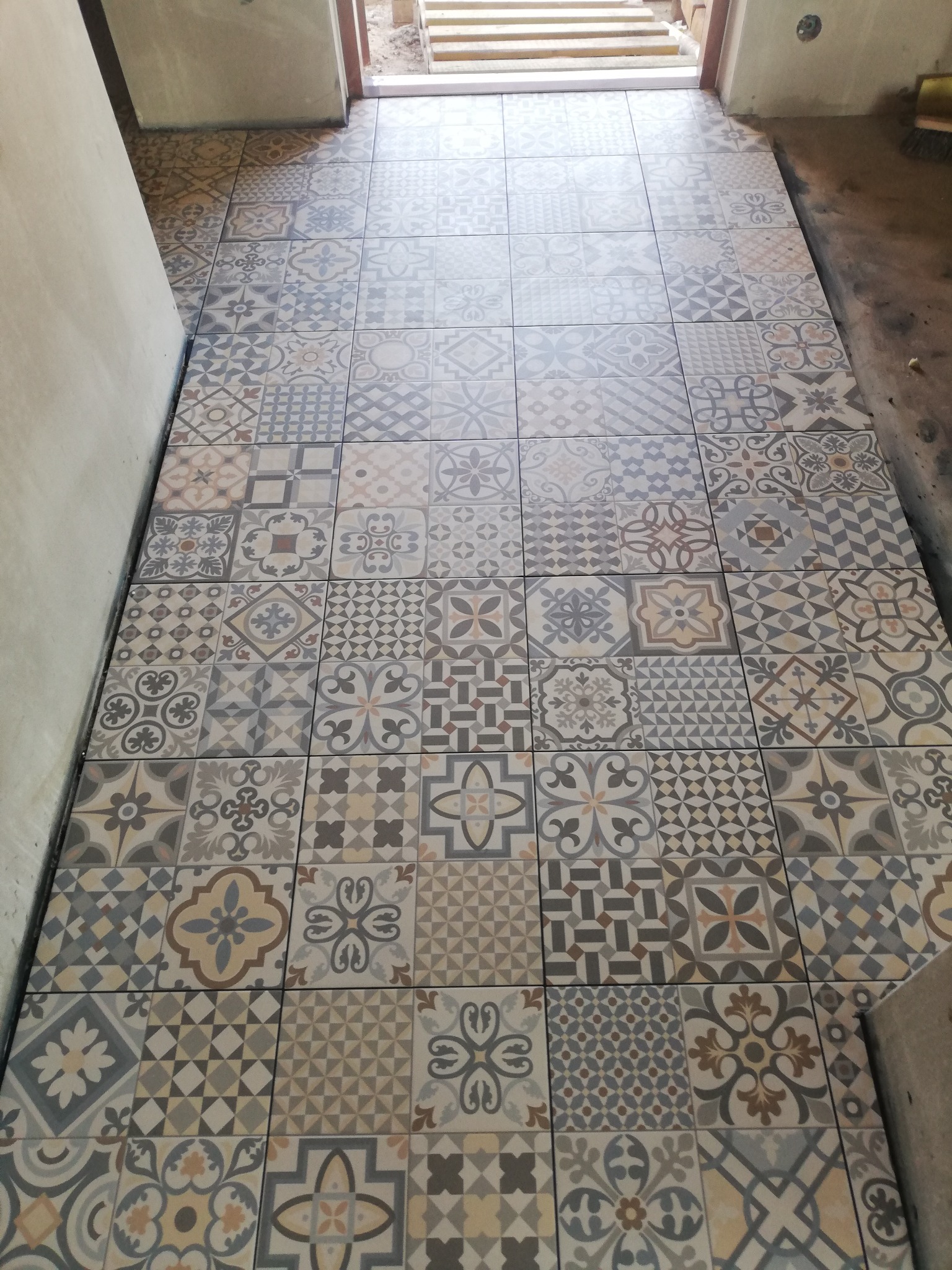 Ułożona podłoga z patchworkowych płytek ceramicznych w odcieniach szarości, beżu i brązu, widok z góry na korytarz w trakcie remontu.