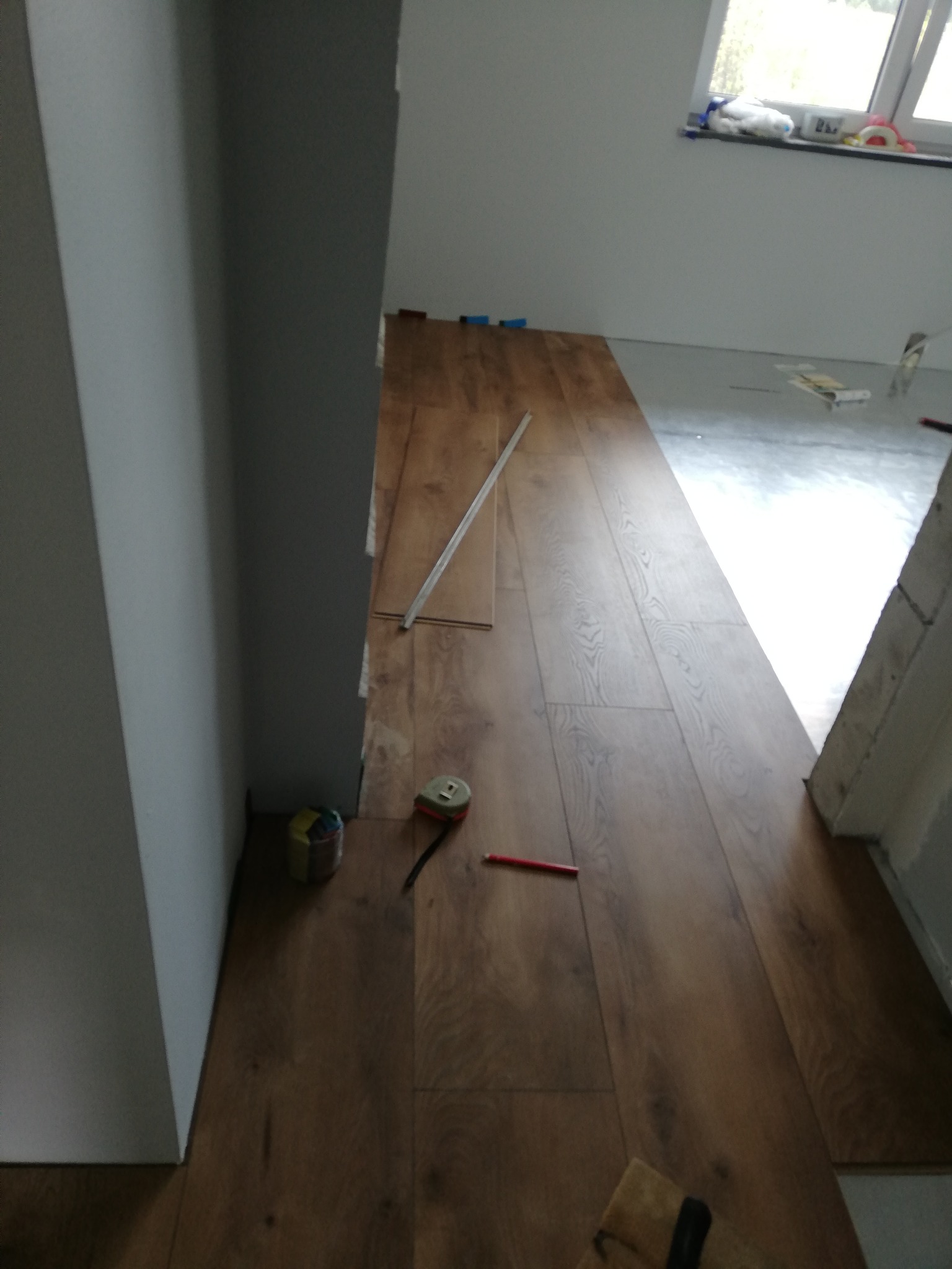 Fragment układania paneli podłogowych w pomieszczeniu, widoczna część ułożonej podłogi, narzędzia pracy: miarka, ołówek, listwa i paczka kolorowych gumek recepturek przy ścianie.