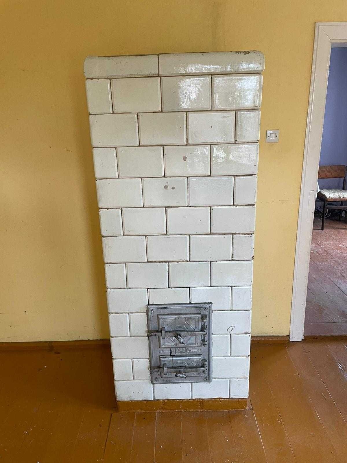 Stary piec kaflowy z metalowymi drzwiczkami na tle żółtej ściany i drewnianej podłogi. Widoczny fragment drzwi do innego pomieszczenia.