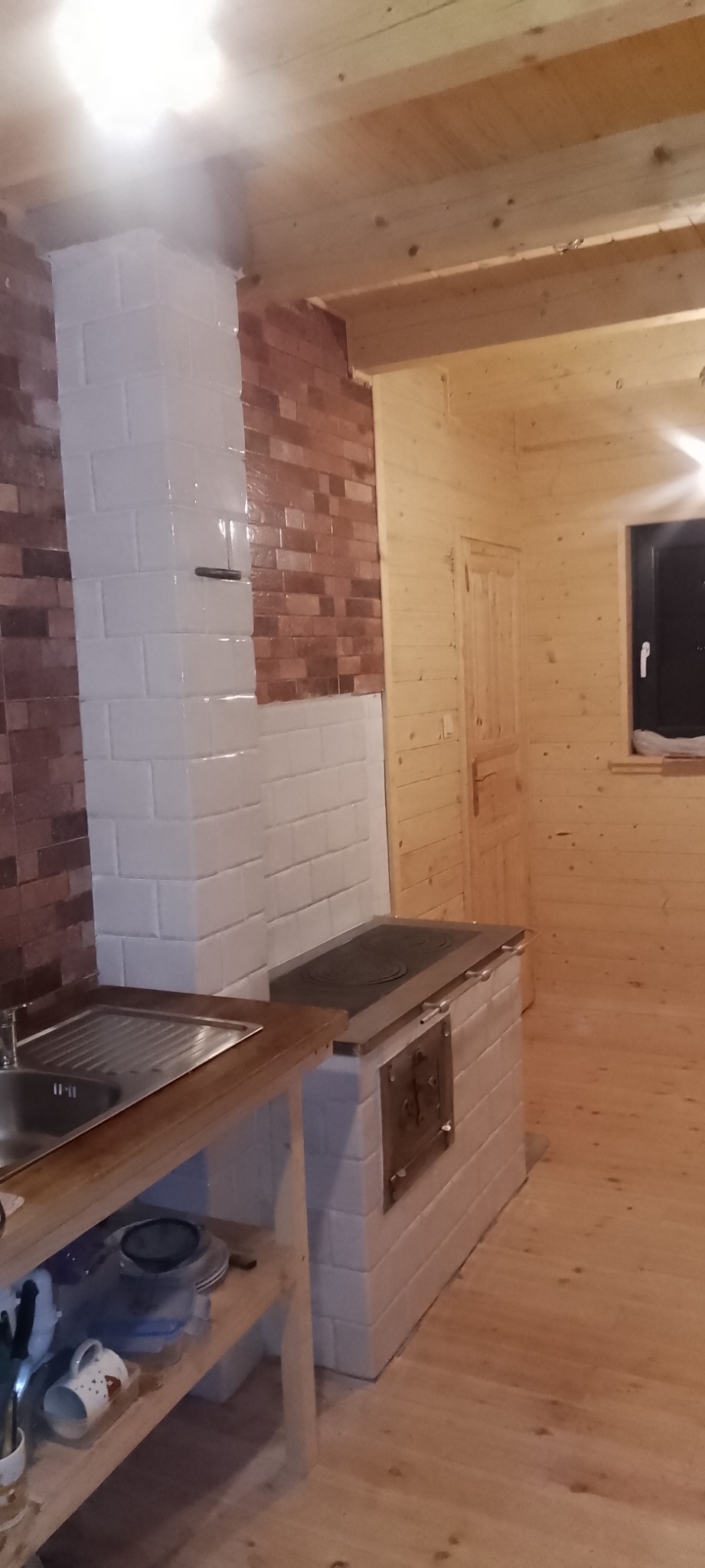 Kuchnia w drewnie z piecem kaflowym i ceglaną ścianą, widok na zlew i blat roboczy.