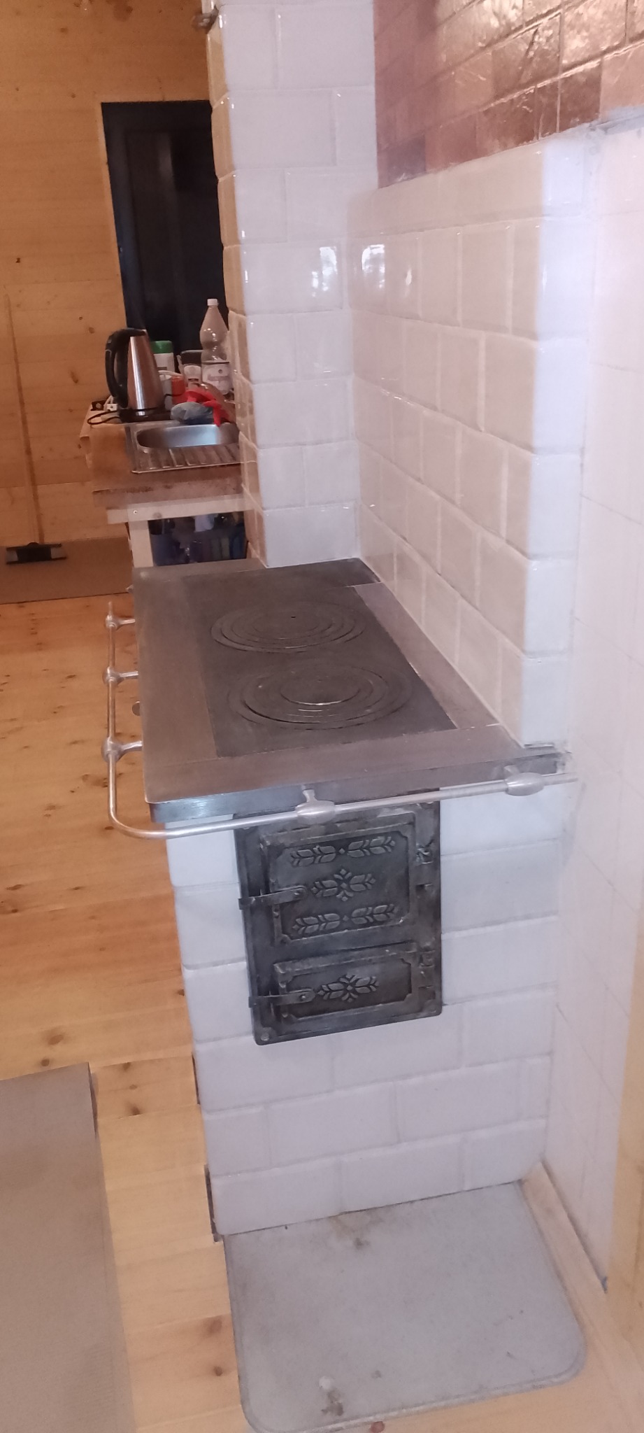 Tradycyjny piec kaflowy z żeliwną płytą grzewczą i ozdobnymi drzwiczkami paleniska, wykończony białymi kaflami w domu w Woli Wodyńskiej.