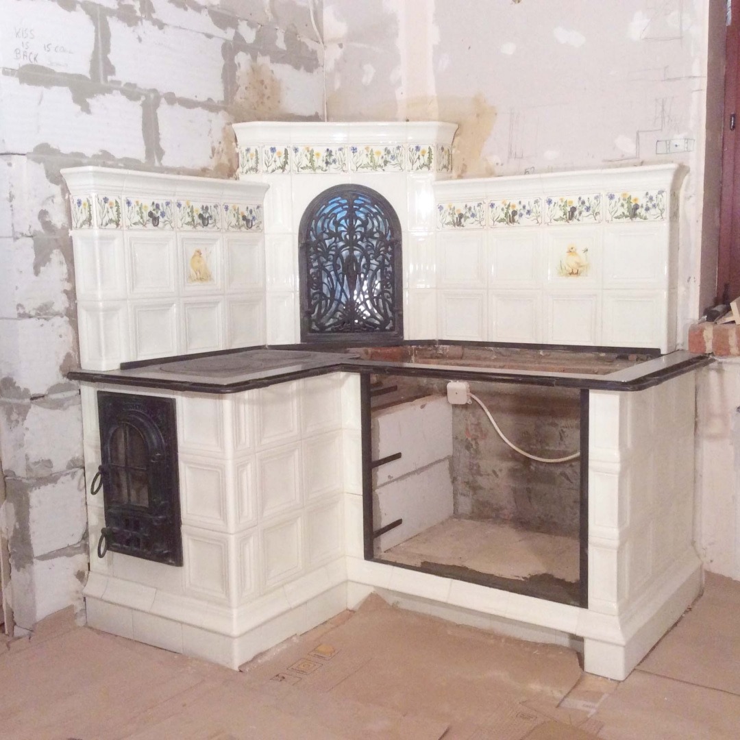 Fragment budowy kaflowego pieca kuchennego z ozdobną, ażurową kratą, białymi kaflami z motywem kwiatowym i zwierzęcym, widoczne niedokończone ściany i podłoga zabezpieczona kartonem.