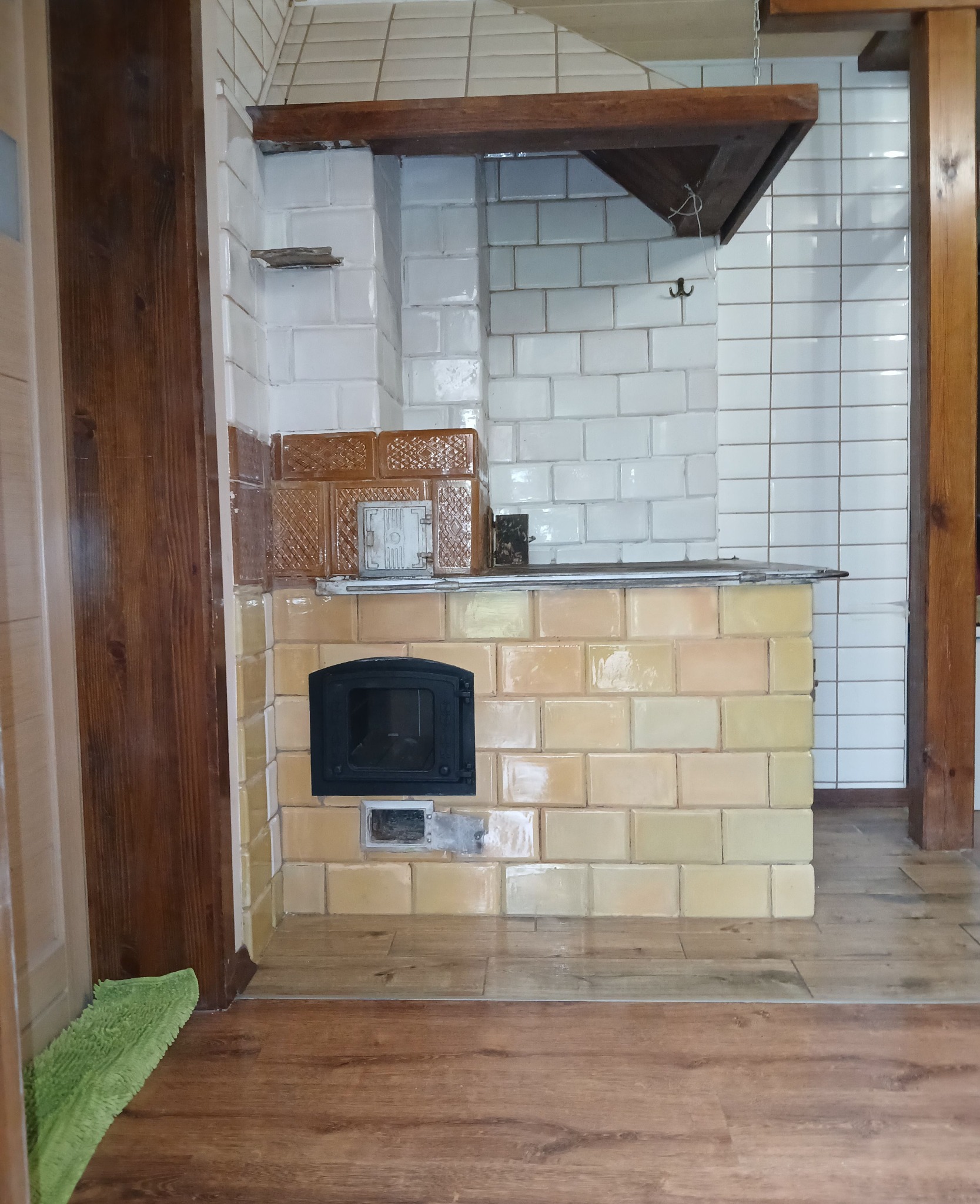 Piec kaflowy z piekarnikiem, obudowany beżowymi i brązowymi kaflami, z drewnianą półką nad paleniskiem i drewnianą konstrukcją nad nim. Widoczna podłoga z paneli.